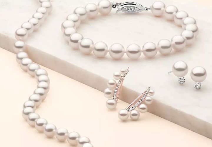 An assortment of white pearl jewelry. A pearl bracelet clasped in white gold and diamond trio, lays atop a marble slab with a pair of pearl and diamond studs and a pair of climbers of 5 graduated pearls on pavé set rose gold curve. A pearl strand drapes over the edge of the slab.